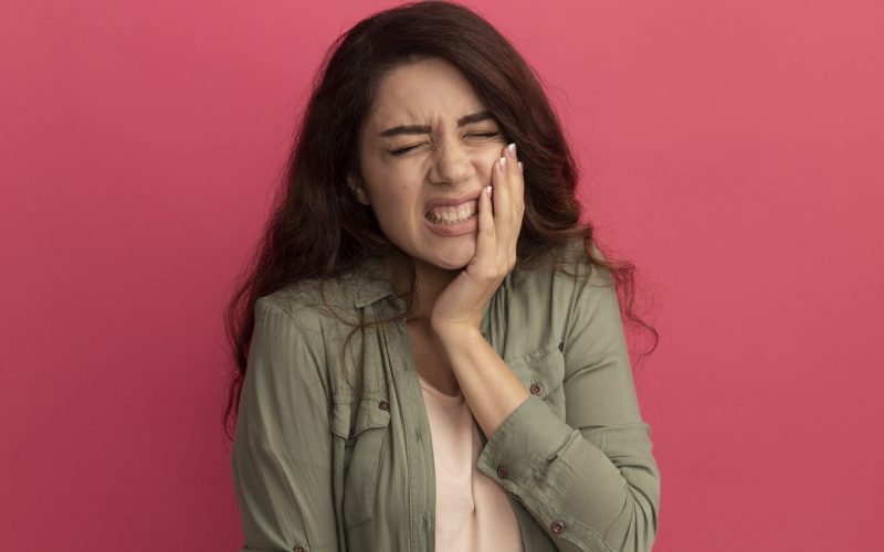 aching young beautiful girl wearing olive green t-shirt putting hand on aching tooth isolated on pink background